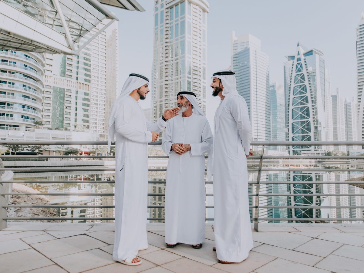 Three Emirati men in conversation on a terrace, high-rise towers behind them